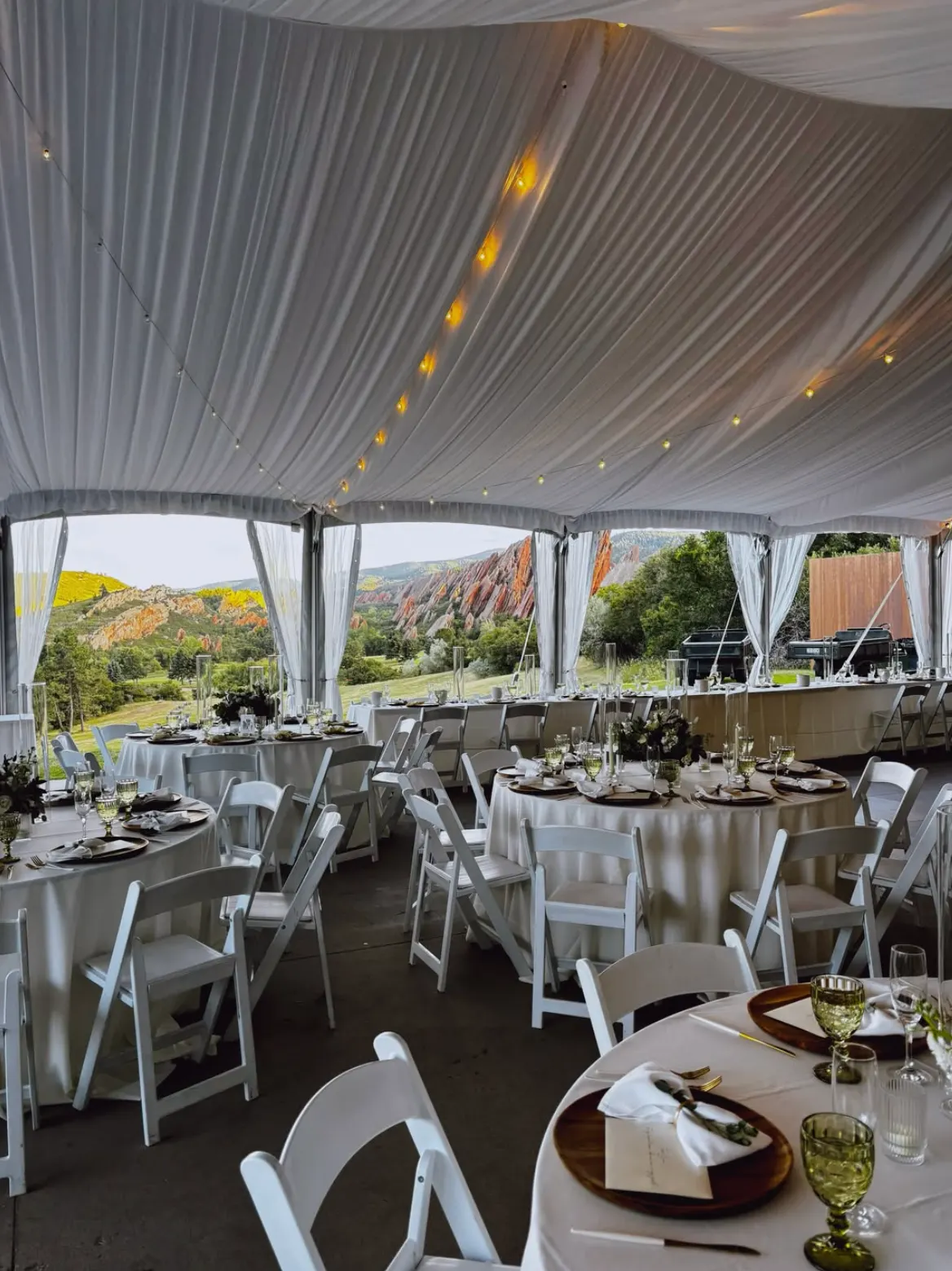 An evening reception tent with round tables set beneath string lights and a red-rock view